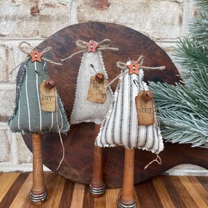 May include: Three handmade fabric Christmas trees with wooden spools for bases. The trees are decorated with burlap ribbon, twine, and small bells. The trees are labeled "Joy", "Merry", and "Noel".