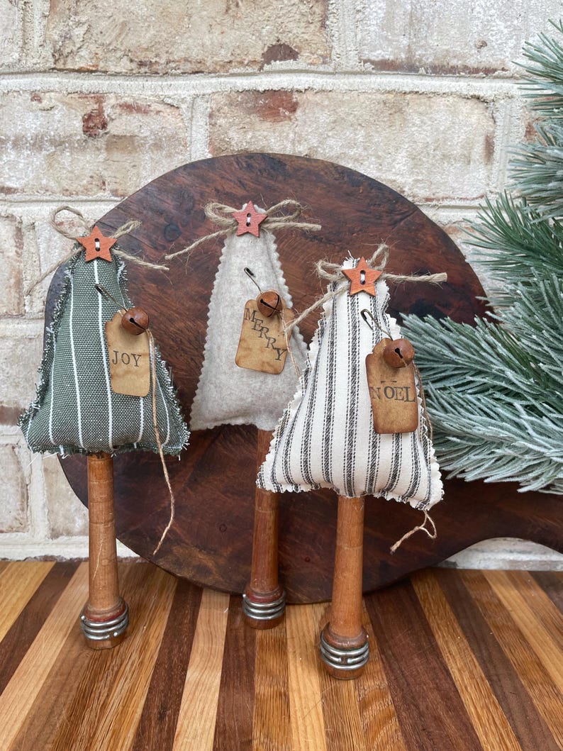 May include: Three handmade fabric Christmas trees with wooden spools for bases. The trees are decorated with burlap ribbon, twine, and small bells. The trees are labeled "Joy", "Merry", and "Noel".