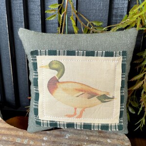 May include: A square sage green fabric pillow featuring a central rectangular patch. The patch displays a printed illustration of a mallard duck with a green head, brown chest, and tan body, framed by a dark green and white plaid fabric border.