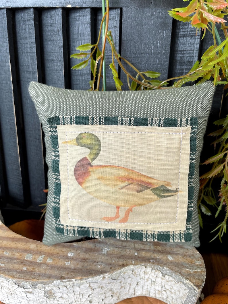 May include: A square sage green fabric pillow featuring a central rectangular patch. The patch displays a printed illustration of a mallard duck with a green head, brown chest, and tan body, framed by a dark green and white plaid fabric border.