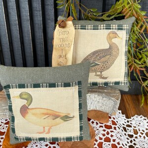 May include: Two square decorative pillows featuring mallard duck illustrations on cream fabric panels with dark green plaid borders. The pillows are made of textured olive green fabric. A wooden tag attached to the back pillow reads INTO THE WOODS I GO.