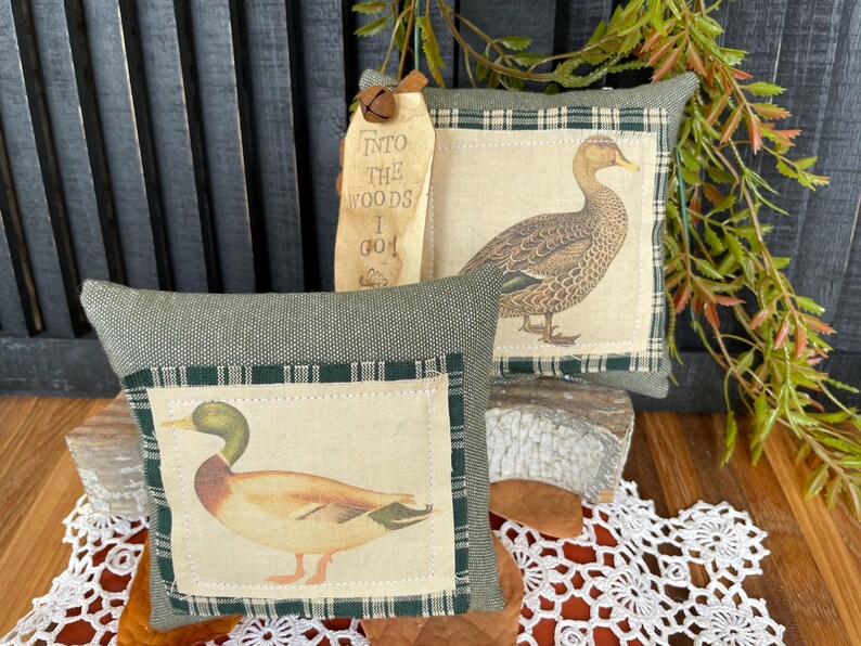 May include: Two square decorative pillows featuring mallard duck illustrations on cream fabric panels with dark green plaid borders. The pillows are made of textured olive green fabric. A wooden tag attached to the back pillow reads INTO THE WOODS I GO.