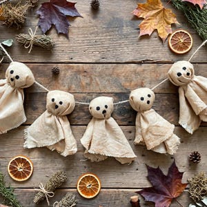 May include: A garland featuring four beige fabric ghost figures strung on a thin cord. Each ghost has a rounded head with two black circular eyes and a small black circular mouth. The fabric bodies have frayed edges and a natural, textured appearance.