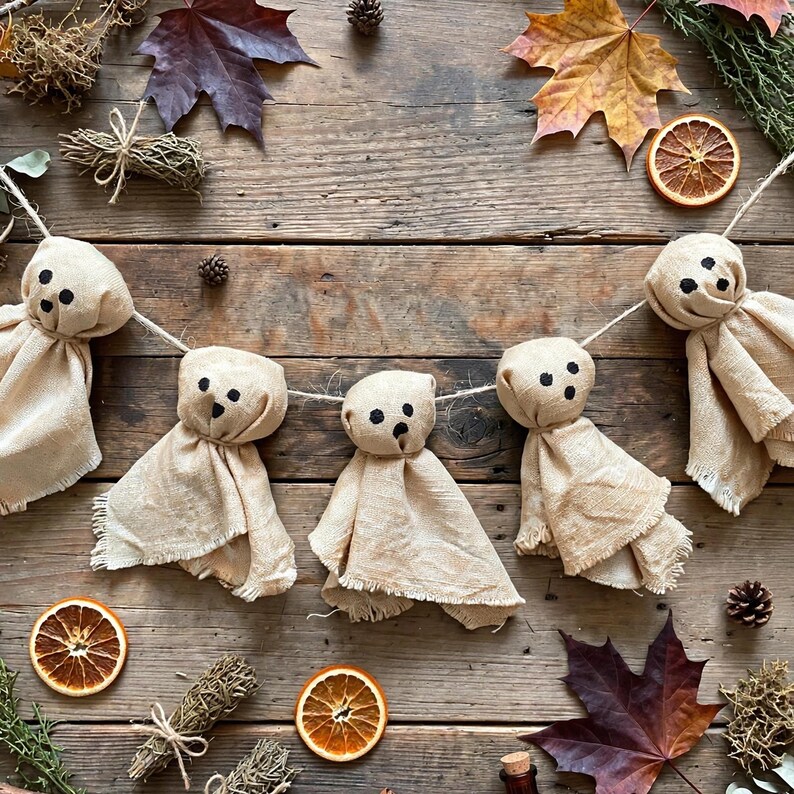 May include: A garland featuring four beige fabric ghost figures strung on a thin cord. Each ghost has a rounded head with two black circular eyes and a small black circular mouth. The fabric bodies have frayed edges and a natural, textured appearance.