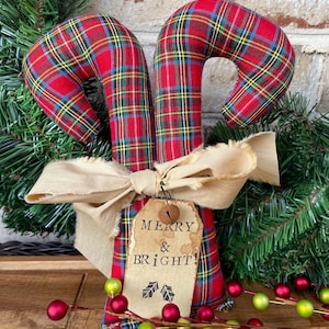 May include: Two fabric candy canes featuring a red, green, yellow, and blue plaid pattern. A beige frayed ribbon is tied around the center, holding a tan paper tag with the printed text MERRY & BRIGHT! and a small metal jingle bell.