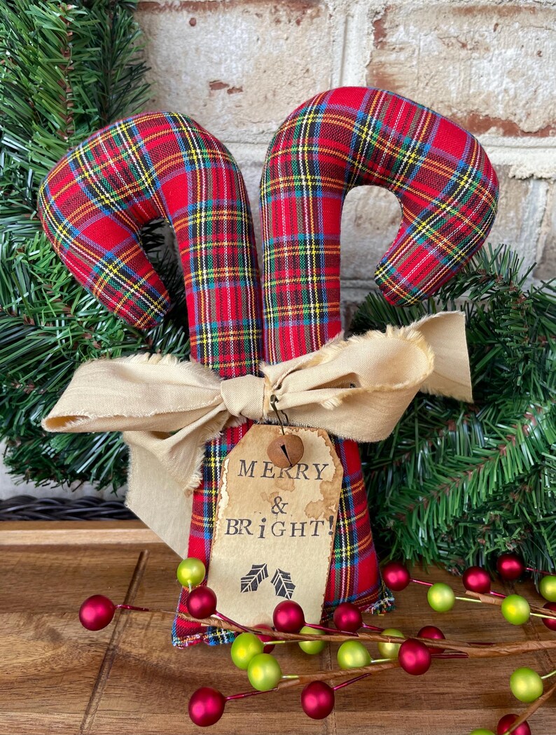 May include: Two fabric candy canes featuring a red, green, yellow, and blue plaid pattern. A beige frayed ribbon is tied around the center, holding a tan paper tag with the printed text MERRY & BRIGHT! and a small metal jingle bell.