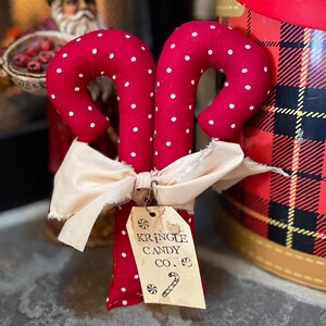 May include: Two red fabric candy canes with white polka dots, tied together with a cream-colored bow. A tag reads "Kringle Candy Co." with a candy cane illustration. Festive holiday decor.