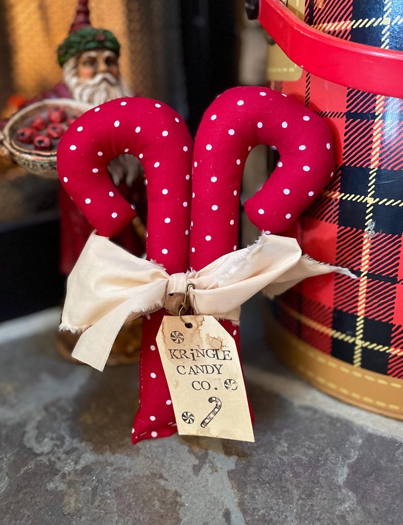 May include: Two red fabric candy canes with white polka dots, tied together with a cream-colored bow. A tag reads "Kringle Candy Co." with a candy cane illustration. Festive holiday decor.