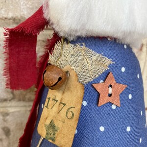 K&ouml;nnte beinhalten: Eine blaue Stoffpuppe mit wei&szlig;en Punkten, einem roten Schal und einer braunen Glocke. Die Puppe hat ein Schild mit der Nummer 1776 und einen Holzstern.