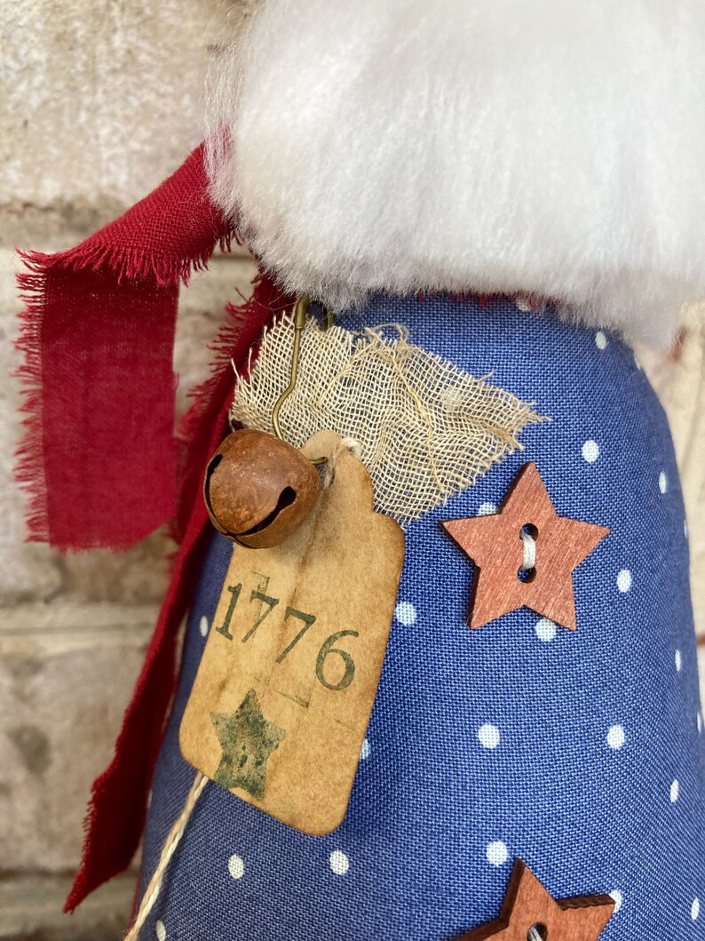 K&ouml;nnte beinhalten: Eine blaue Stoffpuppe mit wei&szlig;en Punkten, einem roten Schal und einer braunen Glocke. Die Puppe hat ein Schild mit der Nummer 1776 und einen Holzstern.