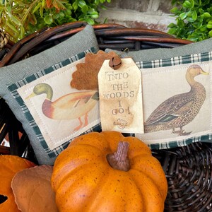 May include: A rectangular green fabric pillow featuring two duck illustrations on cream-colored patches, a brown felt leaf, and a beige paper tag with the text "INTO THE WOODS I GO!" attached by a small metal bell, all nestled in a dark wicker basket.