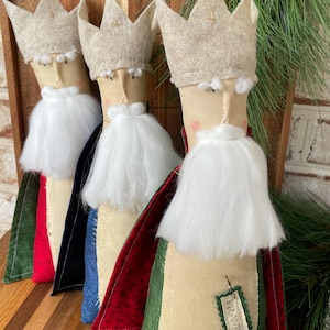 May include: Handmade Christmas ornaments featuring three kings with beige fabric faces, white beards, and felt crowns. Each king wears a different colored cape: red, black, blue, and green. A tag with text is attached to one of the figures.