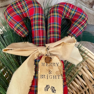 May include: Two fabric candy canes featuring a red, green, yellow, and blue plaid pattern. A beige frayed ribbon is tied around the center, holding a rectangular paper tag with the text MERRY & BRIGHT! and two small leaf illustrations.