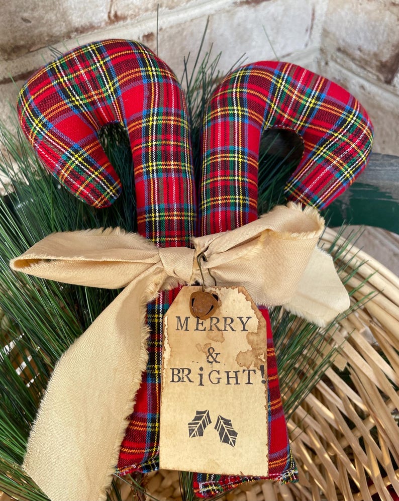 May include: Two fabric candy canes featuring a red, green, yellow, and blue plaid pattern. A beige frayed ribbon is tied around the center, holding a rectangular paper tag with the text MERRY & BRIGHT! and two small leaf illustrations.