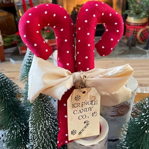 May include: Two red fabric candy canes with white polka dots, tied with a cream-colored bow. A tag reads "Kringle Candy Co." with a candy cane illustration. The candy canes are displayed with small Christmas trees.