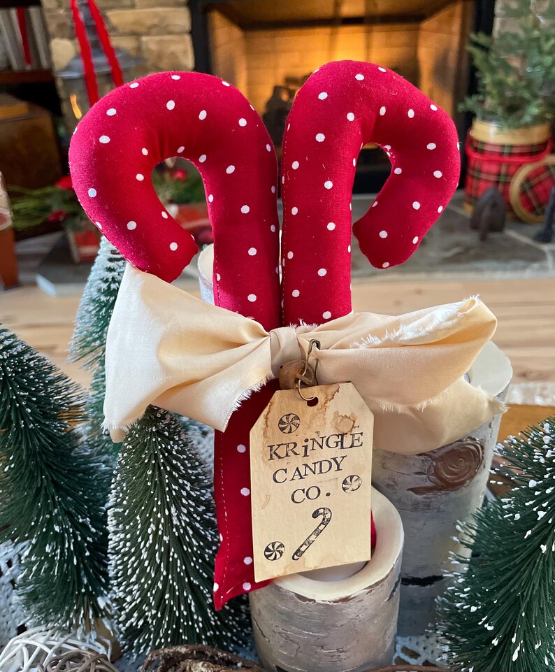 May include: Two red fabric candy canes with white polka dots, tied with a cream-colored bow. A tag reads "Kringle Candy Co." with a candy cane illustration. The candy canes are displayed with small Christmas trees.