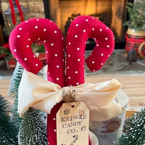 May include: Two red fabric candy canes with white polka dots, tied with a cream-colored bow. A tag reads "Kringle Candy Co." with a candy cane illustration. Festive holiday decor.