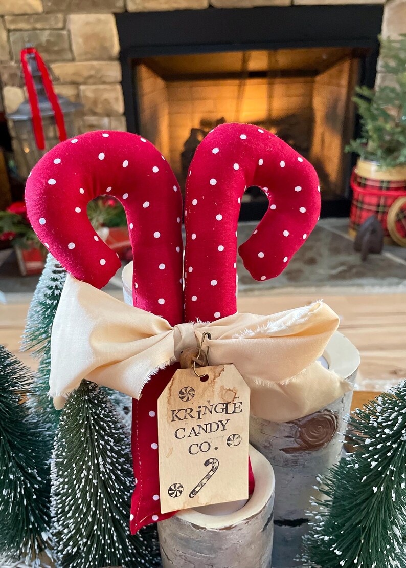 May include: Two red fabric candy canes with white polka dots, tied with a cream-colored bow. A tag reads "Kringle Candy Co." with a candy cane illustration. Festive holiday decor.