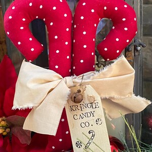 May include: Two red fabric candy canes with white polka dots, tied together with a cream-colored bow. A tag reads "Kringle Candy Co." with a small bell attached. Festive holiday decor.