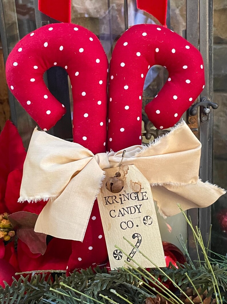May include: Two red fabric candy canes with white polka dots, tied together with a cream-colored bow. A tag reads "Kringle Candy Co." with a small bell attached. Festive holiday decor.