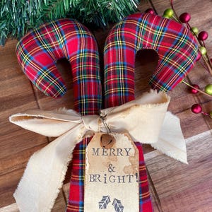 May include: Two fabric candy canes featuring a red, green, and yellow plaid pattern. The canes are tied together with a beige frayed ribbon and a rectangular paper tag that reads MERRY & BRIGHT! with two small holly leaf icons and a metal bell.