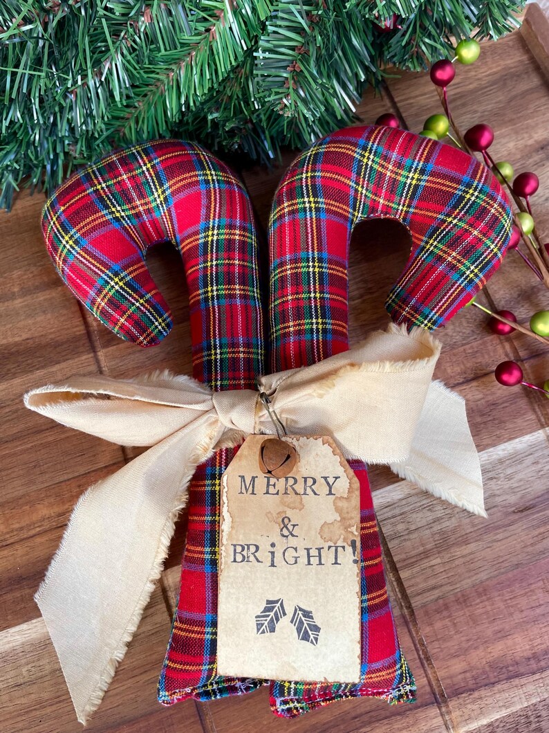 May include: Two fabric candy canes featuring a red, green, and yellow plaid pattern. The canes are tied together with a beige frayed ribbon and a rectangular paper tag that reads MERRY & BRIGHT! with two small holly leaf icons and a metal bell.