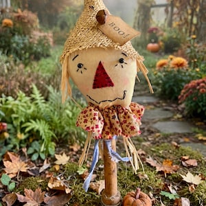 May include: A fabric scarecrow doll with a burlap hat, a red stitched nose, and a tan tag labeled "Boo!" attached with a small acorn. The doll features a floral ruffled collar, a wooden stick base, and a small fabric pumpkin at its feet.