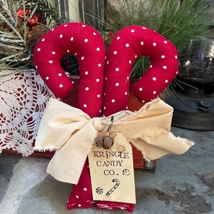 May include: Two red fabric candy canes with white polka dots, tied together with a cream-colored bow. A small tag reads "Kringle Candy Co." with a candy cane illustration. Festive holiday decor.