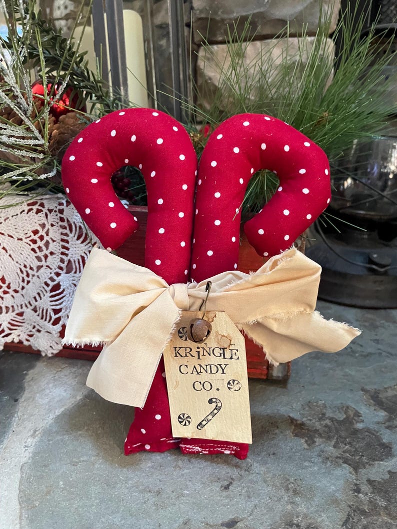 May include: Two red fabric candy canes with white polka dots, tied together with a cream-colored bow. A small tag reads "Kringle Candy Co." with a candy cane illustration. Festive holiday decor.