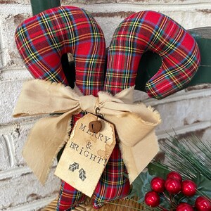 May include: A fabric candy cane ornament featuring a red, green, and yellow plaid pattern. It is tied with a beige frayed ribbon and a tan paper tag that reads MERRY & BRIGHT! with two small holly leaf icons and a small metal jingle bell attached.