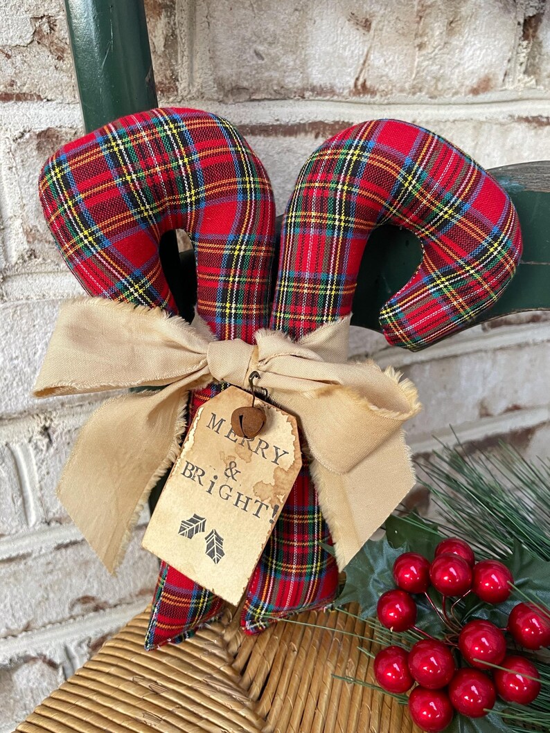 May include: A fabric candy cane ornament featuring a red, green, and yellow plaid pattern. It is tied with a beige frayed ribbon and a tan paper tag that reads MERRY & BRIGHT! with two small holly leaf icons and a small metal jingle bell attached.