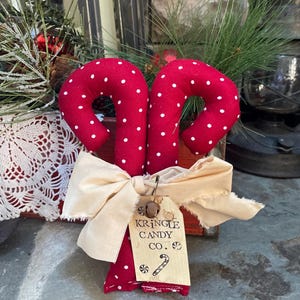 May include: Red fabric candy canes with white polka dots, tied with a cream-colored bow. A tag reads "KRINGLE CANDY CO." with a candy cane illustration. Festive holiday decor.