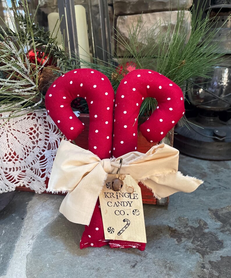 May include: Red fabric candy canes with white polka dots, tied with a cream-colored bow. A tag reads "KRINGLE CANDY CO." with a candy cane illustration. Festive holiday decor.