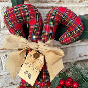 May include: Two fabric candy canes featuring a red, green, yellow, and blue plaid pattern. A beige frayed ribbon is tied around the center, holding a tan paper tag with the text MERRY & BRIGHT! and two small leaf icons, topped with a metal bell.