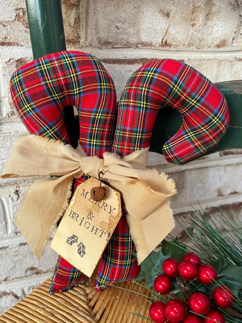 May include: Two fabric candy canes featuring a red, green, yellow, and blue plaid pattern. A beige frayed ribbon is tied around the center, holding a tan paper tag with the text MERRY & BRIGHT! and two small leaf icons, topped with a metal bell.