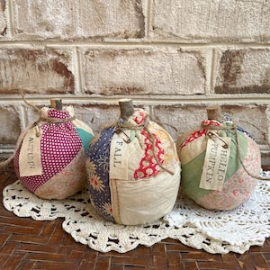 May include: Three patchwork fabric pumpkins with different fall-themed labels: "Autumn", "Fall", and "Hello Pumpkin". The pumpkins are sitting on a white crocheted doily on a woven brown surface.