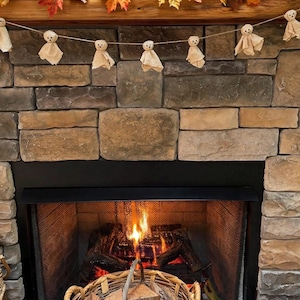 May include: A garland of small, beige fabric ghost figures with simple drawn faces, strung on twine and hung across a stone fireplace. Above the ghosts, a decorative autumn garland features orange, yellow, and brown maple leaves and berries.