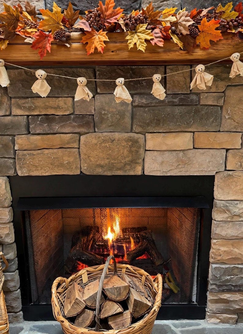 May include: A garland of small, beige fabric ghost figures with simple drawn faces, strung on twine and hung across a stone fireplace. Above the ghosts, a decorative autumn garland features orange, yellow, and brown maple leaves and berries.