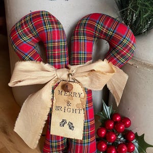 May include: Two fabric candy canes featuring a red, yellow, and blue plaid pattern. A beige frayed ribbon is tied around the center, holding a tan paper tag with the text MERRY & BRIGHT! and two small holly leaf icons, topped with a metal bell.