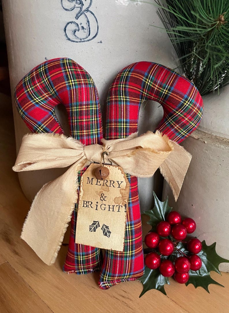 May include: Two fabric candy canes featuring a red, yellow, and blue plaid pattern. A beige frayed ribbon is tied around the center, holding a tan paper tag with the text MERRY & BRIGHT! and two small holly leaf icons, topped with a metal bell.