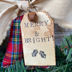 May include: A red, blue, and yellow plaid fabric ornament featuring a beige frayed ribbon bow and a metal safety pin holding a rectangular paper tag. The tag has stained edges and the black text MERRY & BRIGHT! with two holly leaf icons.