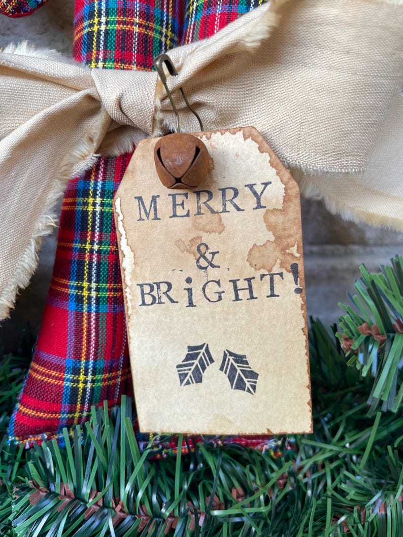 May include: A red, blue, and yellow plaid fabric ornament featuring a beige frayed ribbon bow and a metal safety pin holding a rectangular paper tag. The tag has stained edges and the black text MERRY & BRIGHT! with two holly leaf icons.