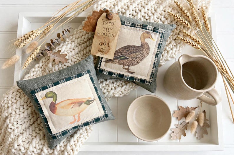 May include: Two square gray fabric pillows featuring a dark green plaid border and a central patch with a duck illustration. One pillow has a tan tag attached with the text "INTO THE WOODS I GO". Also includes a ceramic pitcher and a small bowl.