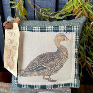May include: A square green fabric pillow featuring a central patch with a detailed illustration of a brown duck. The patch is framed by a green and white plaid border. A tan tag attached with a metal bell reads, INTO THE WOODS I GO!