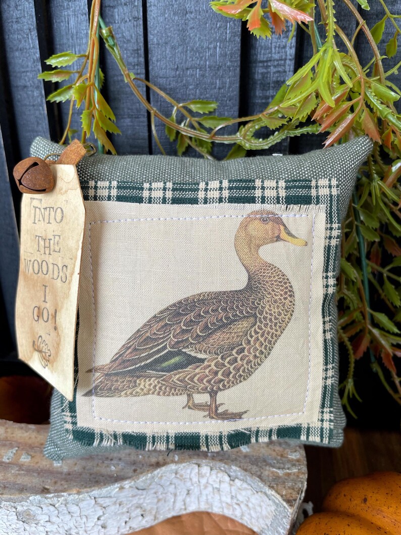 May include: A square green fabric pillow featuring a central patch with a detailed illustration of a brown duck. The patch is framed by a green and white plaid border. A tan tag attached with a metal bell reads, INTO THE WOODS I GO!