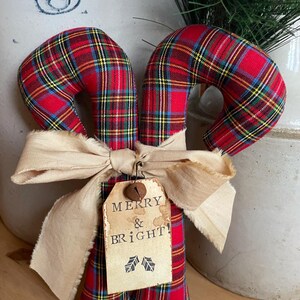 May include: Two fabric candy canes featuring a red, yellow, and blue plaid pattern. A beige frayed ribbon is tied around the center, holding a rectangular paper tag with the text MERRY & BRIGHT! and two small holly leaf illustrations.