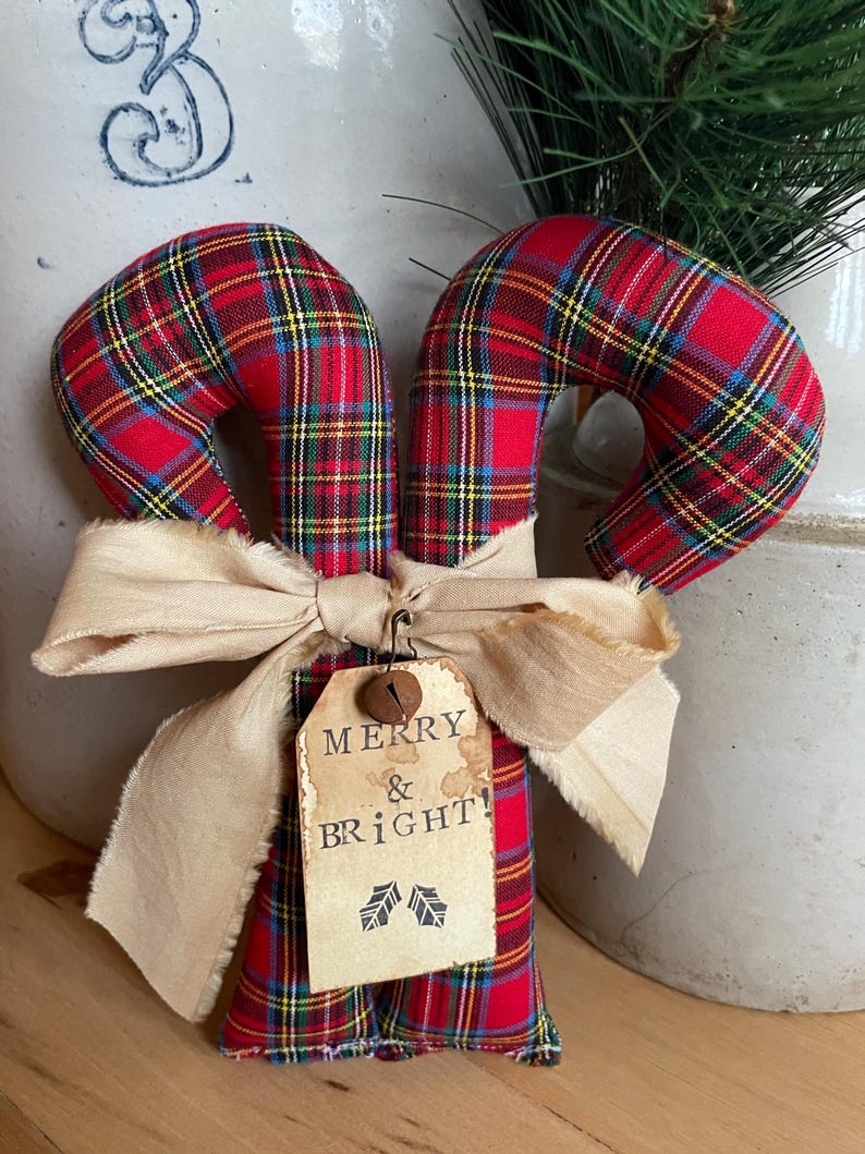 May include: Two fabric candy canes featuring a red, yellow, and blue plaid pattern. A beige frayed ribbon is tied around the center, holding a rectangular paper tag with the text MERRY & BRIGHT! and two small holly leaf illustrations.