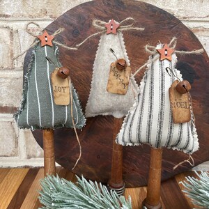May include: Three handmade fabric Christmas tree ornaments with wooden dowel bases. The trees are decorated with burlap ribbon, a star, and a bell. The fabric is striped in black and white, black and green, and white. The ornaments have tags that read "Joy", "Merry", and "Noel".