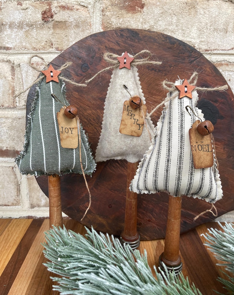 May include: Three handmade fabric Christmas tree ornaments with wooden dowel bases. The trees are decorated with burlap ribbon, a star, and a bell. The fabric is striped in black and white, black and green, and white. The ornaments have tags that read "Joy", "Merry", and "Noel".