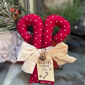 May include: Two red fabric candy canes with white polka dots are tied together with a cream-colored bow. A small tag reads "KRINGLE CANDY CO." with a candy cane illustration. Festive holiday decor.